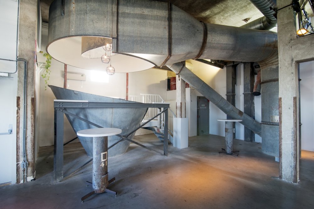 Inrichting met hergebruikte fabrieksonderdelen: statafels gemaakt van oude stalen buizen, rond de originele trechters en leidingen van het pand | maatwerk van Luc en Roel, Rotterdam