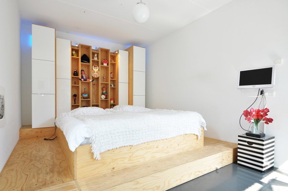 Slaapkamer met een bed op een houten podium: brede treden eromheen en een wand van kasten en open vakken als hoofdeinde | maatwerk van Luc en Roel, Rotterdam