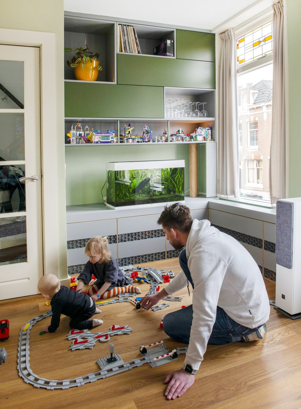 Wandmeubel om het raam heen, met groene en grijze kastjes, open vakken en een doorlopende vensterbank, deels gemaakt van hergebruikt materiaal | maatwerk van Luc en Roel, Rotterdam