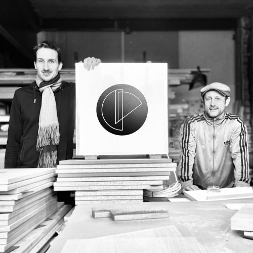 Luc en Roel in hun werkplaats in Rotterdam, met het logo tussen stapels plaatmateriaal